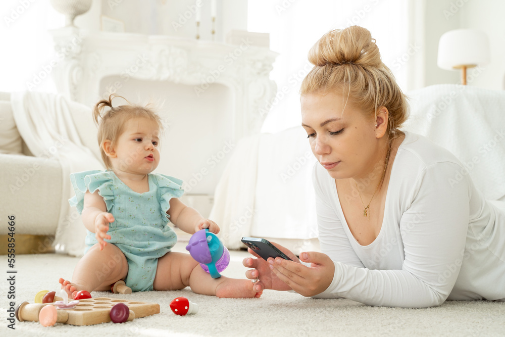 Young mom using smartphone while playing with baby. Lack of live communication, Addiction on gadgets. Social media, watch video, Woman search for products for children in Internet. Young mom using smartphone while playing with baby. Lack of live communication, Addiction on gadgets. Social media, watch video, Woman search for products for children in Internet.