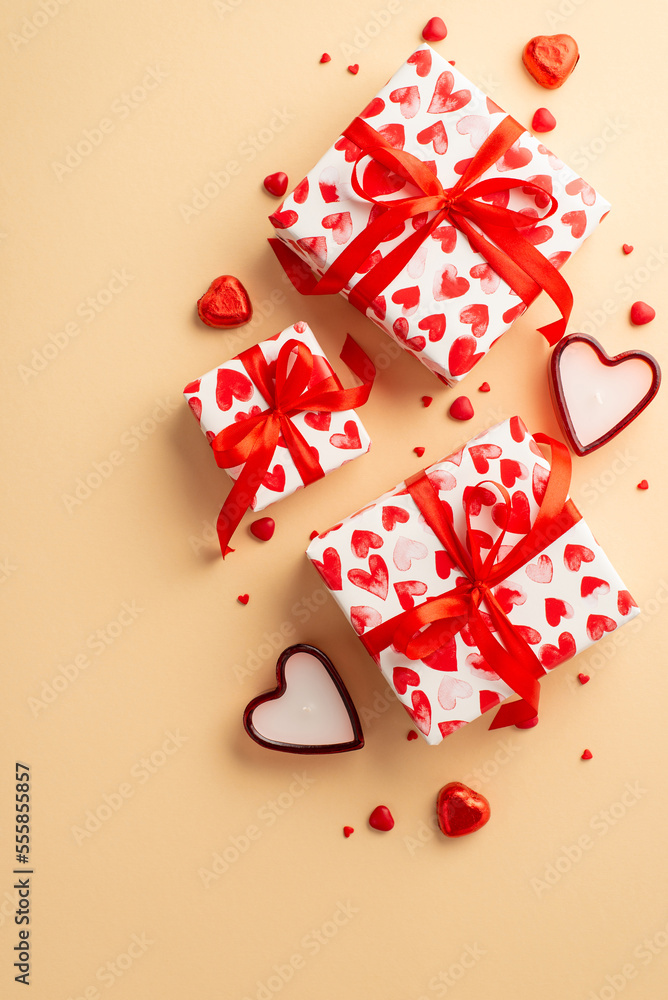 Saint Valentine's Day concept. Top view vertical photo of gift boxes with ribbon bows heart shaped candies candles and sprinkles on isolated light beige background Saint Valentine's Day concept. Top view vertical photo of gift boxes with ribbon bows heart shaped candies candles and sprinkles on isolated light beige background