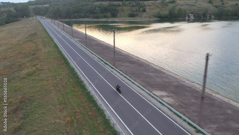 Aerial view to biker riding on modern sport motorbike at road near lake. Motorcyclist racing his motorcycle at dam route on summer day. Man drive bike during trip. Concept of freedom and adventure Aerial view to biker riding on modern sport motorbike at road near lake. Motorcyclist racing his motorcycle at dam route on summer day. Man drive bike during trip. Concept of freedom and adventure
