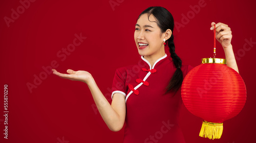 Smiling young asian woman in red traditional chinese dress holding red paper lantern with hand pointing on red background. Chinese New Year concept. photo