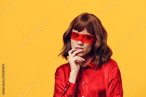 a sweet, thoughtful woman stands in a stylish red shirt and red glasses on a yellow background, thinking and holding her hand near her face. Horizontal Studio Photography © Tatiana