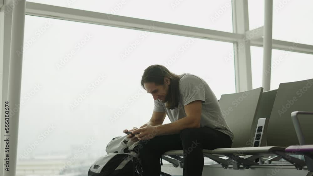 Young backpacker playing a mobile phone videogame sit on an airport while waiting for his departure time Young backpacker playing a mobile phone videogame sit on an airport while waiting for his departure time