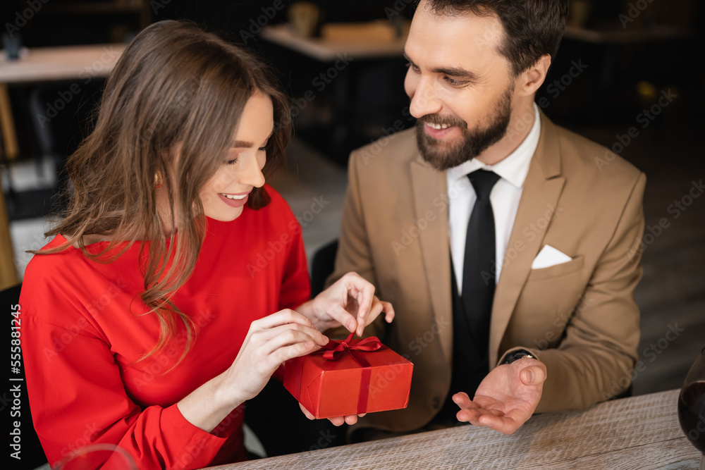 bearded man in formal wear holding present while happy woman in red dress pulling ribbon on valentines day bearded man in formal wear holding present while happy woman in red dress pulling ribbon on valentines day