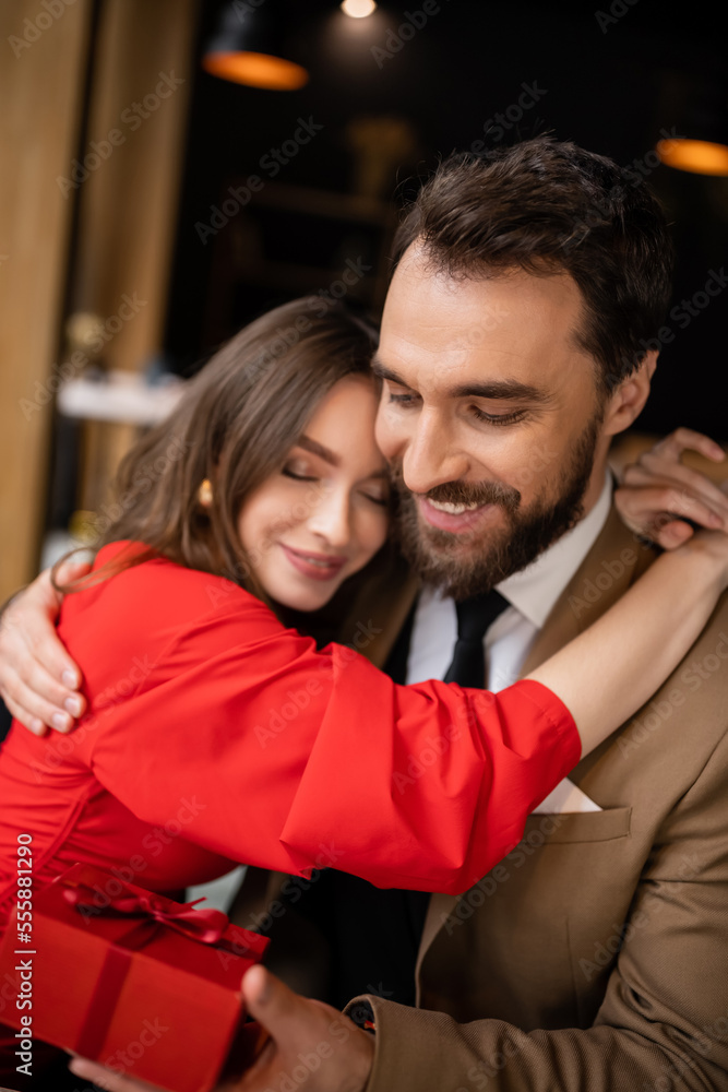 happy woman in red dress hugging bearded boyfriend in formal wear holding present on valentines day happy woman in red dress hugging bearded boyfriend in formal wear holding present on valentines day