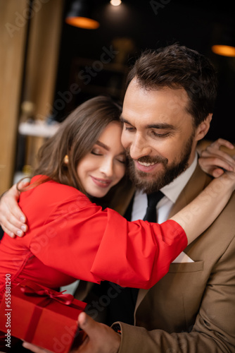 happy woman in red dress hugging bearded boyfriend in formal wear holding present on valentines day