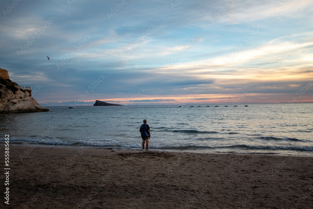Hombre de mediana edad de raza caucásica pensando en solitario sobre el amor en la orilla de la playa de Benidorm con la isla al fondo al atardecer.