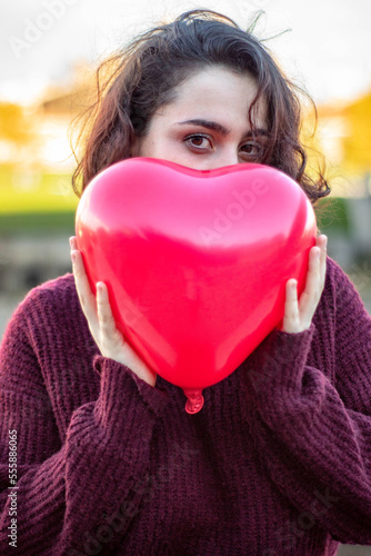 Slika na platnu Mujer joven de raza caucásica con el pelo corto escondiéndose detrás de un globo rojo con forma de corazón que le han regalado por el día de San Valentín