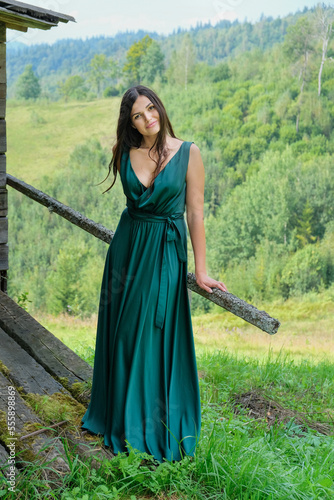 Pretty brunette girl standing at old house in the carpathians. Young romantic woman in long, green, stin dress on the mountainside.
