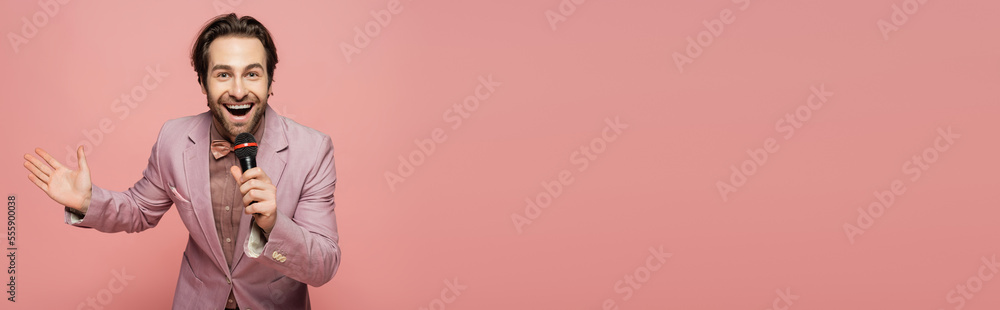Stylish host of event holding microphone and waving hand at camera isolated on pink, banner Stylish host of event holding microphone and waving hand at camera isolated on pink, banner