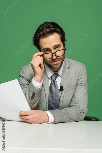 broadcaster in suit adjusting eyeglasses and holding blank paper isolated on green