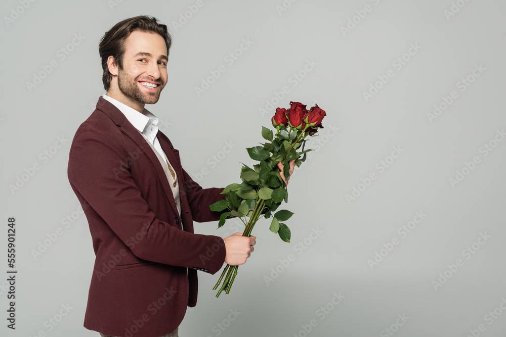 cheerful showman in blazer holding red roses and looking at camera isolated on grey cheerful showman in blazer holding red roses and looking at camera isolated on grey