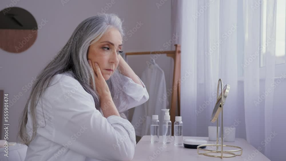 Caucasian old woman looking at mirror reflection touching face gray hair enjoy plastic surgery spa healthy nutrition procedure 50s aged lady female model granny anti age creme cosmetics pampering