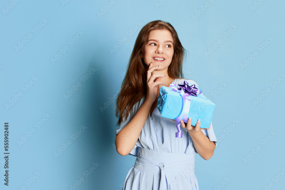 Portrait of young girl in cute tender dress posing over blue background. Birthday celebration. Gifts, presents Portrait of young girl in cute tender dress posing over blue background. Birthday celebration. Gifts, presents
