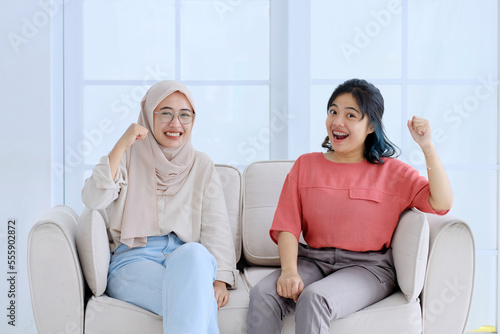 Two beautiful women sitting on sofa raising hands happily looking at camera