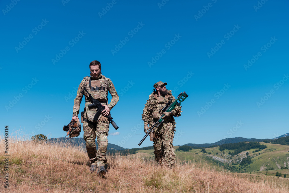 A sniper team squad of soldiers is going undercover. Sniper assistant and team leader walking and aiming in nature with yellow grass and blue sky. Tactical camouflage uniform. A sniper team squad of soldiers is going undercover. Sniper assistant and team leader walking and aiming in nature with yellow grass and blue sky. Tactical camouflage uniform.