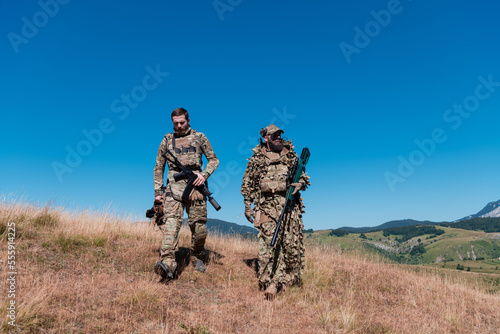 A sniper team squad of soldiers is going undercover. Sniper assistant and team leader walking and aiming in nature with yellow grass and blue sky. Tactical camouflage uniform.