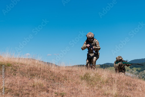 A sniper team squad of soldiers is going undercover. Sniper assistant and team leader walking and aiming in nature with yellow grass and blue sky. Tactical camouflage uniform.