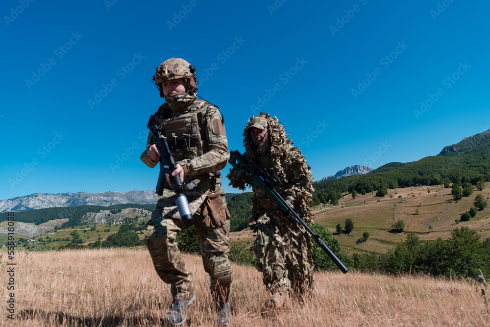 A sniper team squad of soldiers is going undercover. Sniper assistant and team leader walking and aiming in nature with yellow grass and blue sky. Tactical camouflage uniform. A sniper team squad of soldiers is going undercover. Sniper assistant and team leader walking and aiming in nature with yellow grass and blue sky. Tactical camouflage uniform.