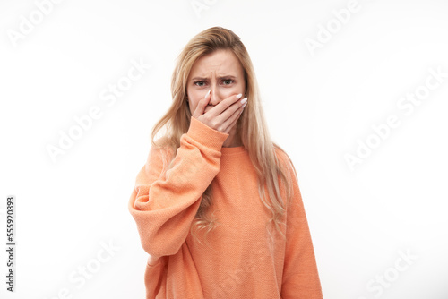 Young blonde woman holding nose to avoid disgusted smell, pinches nose and mouth with fingers and holding breath isolated on white background