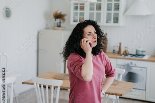 Young curly Italian woman talking by phone in lilac sweatshirt confused by news. Caucasian pretty housewife at kitchen talking using smartphone. Serious arabic woman shocked  upset. Frustrations