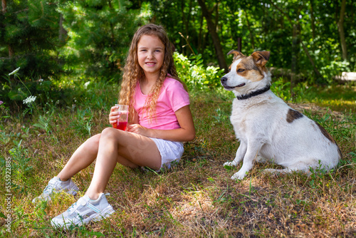 A beautiful and sweet girl sits on the grass with her beloved dog and drinks juice. A happy and smiling child walks in nature with his beloved pet in the summer in the park and drinks soda.