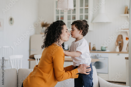 Curly Spanish beautiful mom in orange blouse giving son cookie sitting at desk at kitchen. Caucasian young woman hugs her little curly cheerful son in white t-shirt at home, Little boy kissing mom.