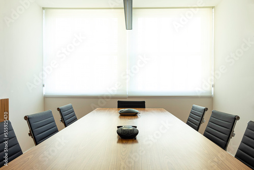 Mesa de reunião de madeira sem ninguém, com equipamentos de comunicação e cadeiras de couro pretas ao seu redor, e ao fundo uma janela com cortinas na cor branca. photo