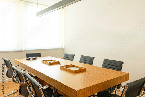 Close da mesa de reunião classica com papeleiras e aparelho de conferência, e ao redor cadeiras de couro preto ao fundo paredes e janelas com cortinas brancas. 