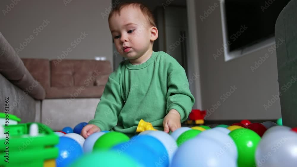 Cute Caucasian child sitting on the floor among the toys. Kid drops one toy and reaches for another. Low angle view. Cute Caucasian child sitting on the floor among the toys. Kid drops one toy and reaches for another. Low angle view.