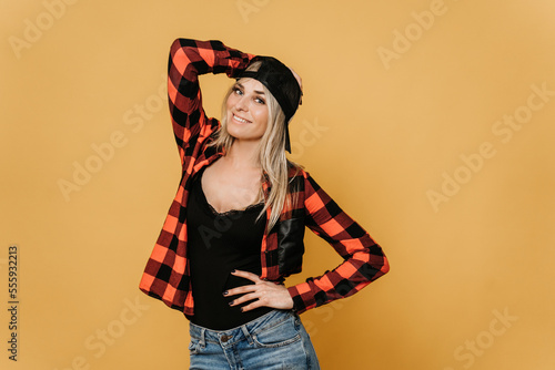 Sexy blonde in baseball cap and checked shirt , broad smiling, posing over yellow background with a copy space for your ad. She is ready to celebrate. Happiness concept.