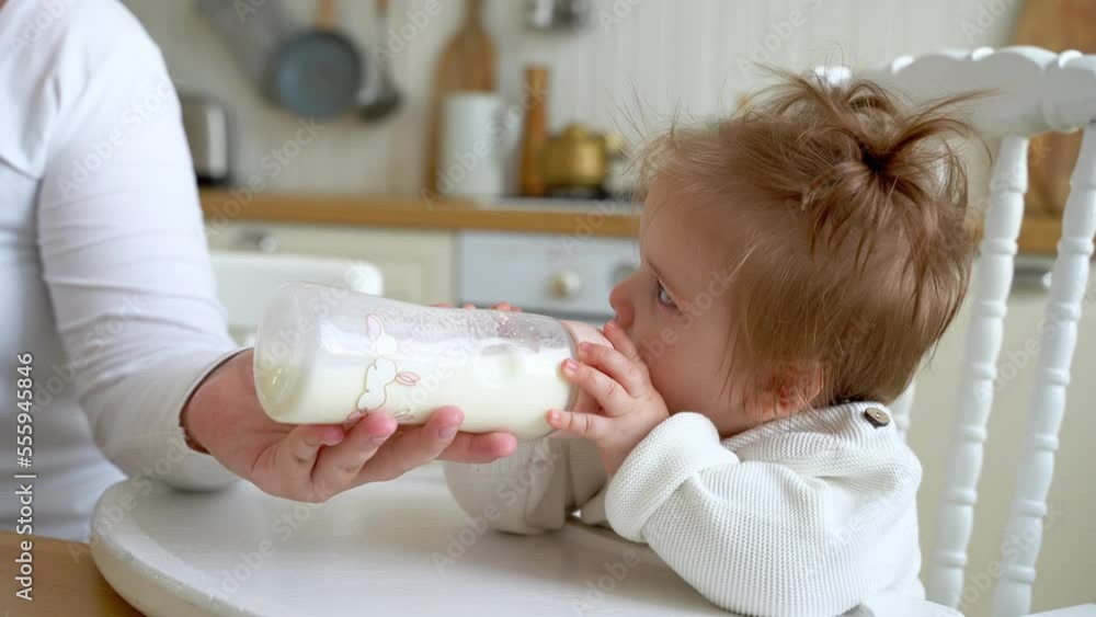 Baby girl drinks powered milk formula from a bottle sitting in high chair at home kitchen. Mother feeding child. Children liquid food. Concept of artificial or breastmilk nourishment. Baby girl drinks powered milk formula from a bottle sitting in high chair at home kitchen. Mother feeding child. Children liquid food. Concept of artificial or breastmilk nourishment.