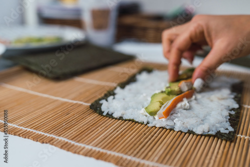 Hands of an unrecognizable woman using her fingers to place a piece of surimi on the sheet of nori seaweed she is using to prepare homemade sushi on her kitchen table.