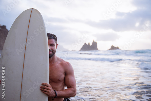 Smiling surfer covers half of his face with surfboard, extreme sports and happiness concept.