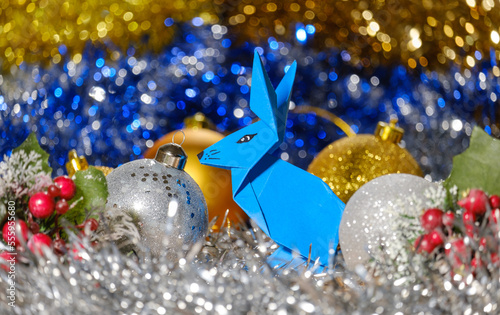 paper blue origami rabbit and Christmas balls on a background of shiny tinsel and bokeh