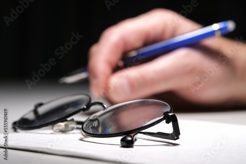 Background, blur, out of focus, bokeh. Glasses in the foreground. In the background, a man is holding a pen between his index and middle fingers, about to write a text.