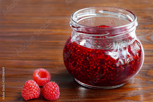 Jar of raspberry jam and fresh raspberries on wooden background, space for text. Opened glass jar of red sweet jam and three fresh berries close-up, copy space, wooden surface