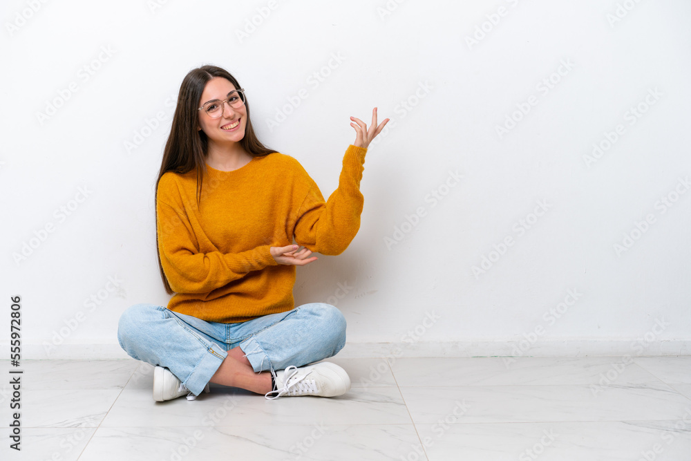 Young girl sitting on the floor isolated on white background extending hands to the side for inviting to come Young girl sitting on the floor isolated on white background extending hands to the side for inviting to come