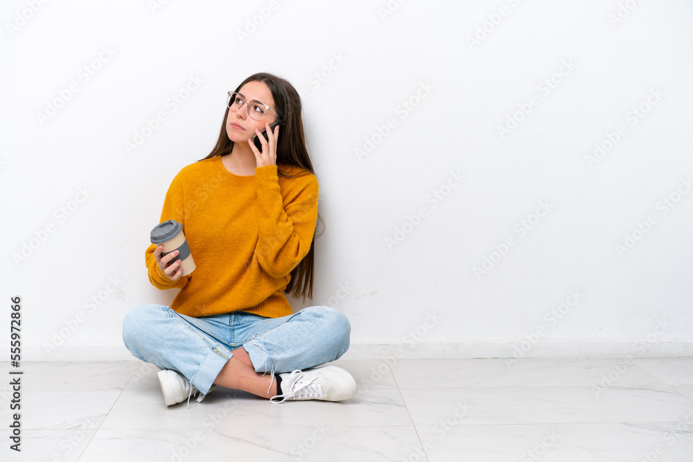 Young girl sitting on the floor isolated on white background holding coffee to take away and a mobile Young girl sitting on the floor isolated on white background holding coffee to take away and a mobile