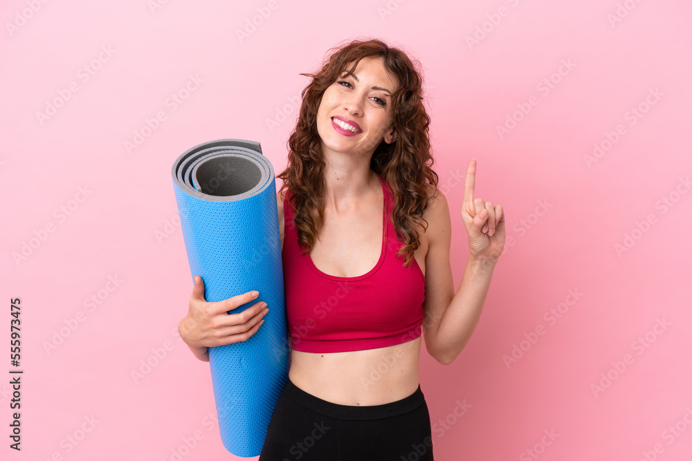Young sport woman going to yoga classes while holding a mat isolated on pink background showing and lifting a finger in sign of the best Young sport woman going to yoga classes while holding a mat isolated on pink background showing and lifting a finger in sign of the best