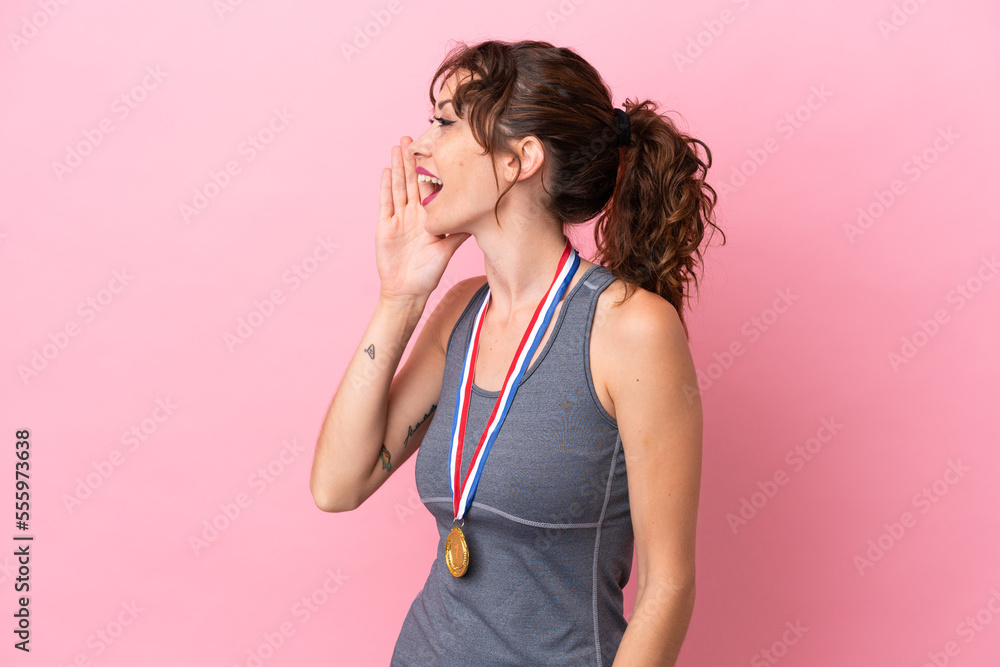 Young caucasian woman with medals isolated on pink background shouting with mouth wide open to the lateral Young caucasian woman with medals isolated on pink background shouting with mouth wide open to the lateral
