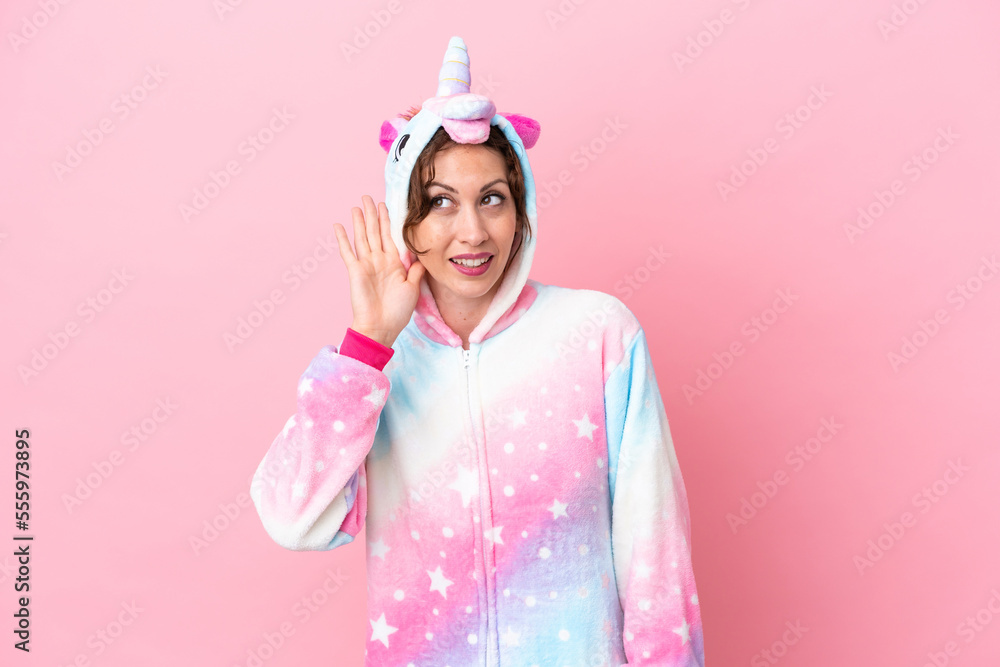 Young caucasian woman with unicorn pajamas isolated on pink background listening to something by putting hand on the ear Young caucasian woman with unicorn pajamas isolated on pink background listening to something by putting hand on the ear