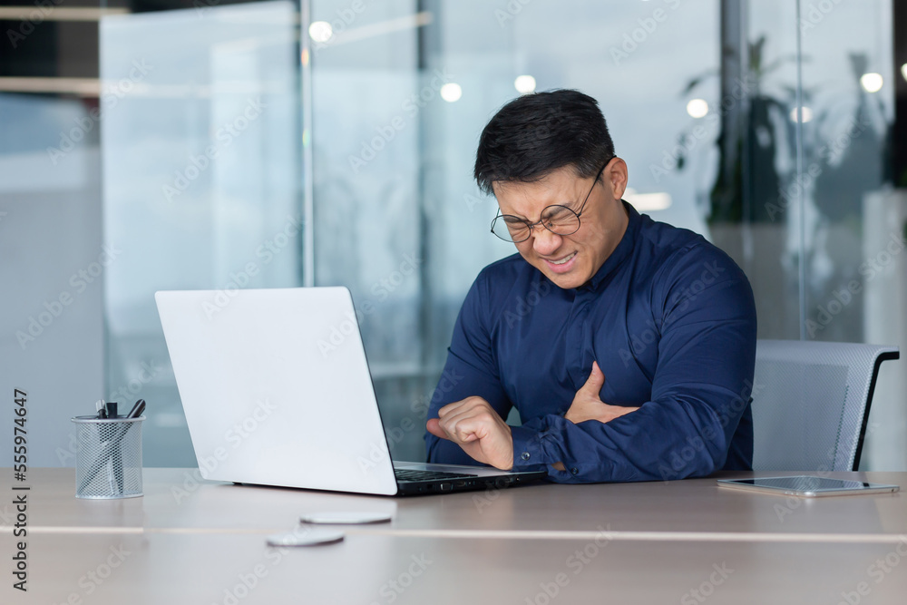 Heart attack at work. A young Asian man is sitting at a desk in the office, holding his heart. A stroke, holding his chest. grimaced, feels severe pain, needs help.