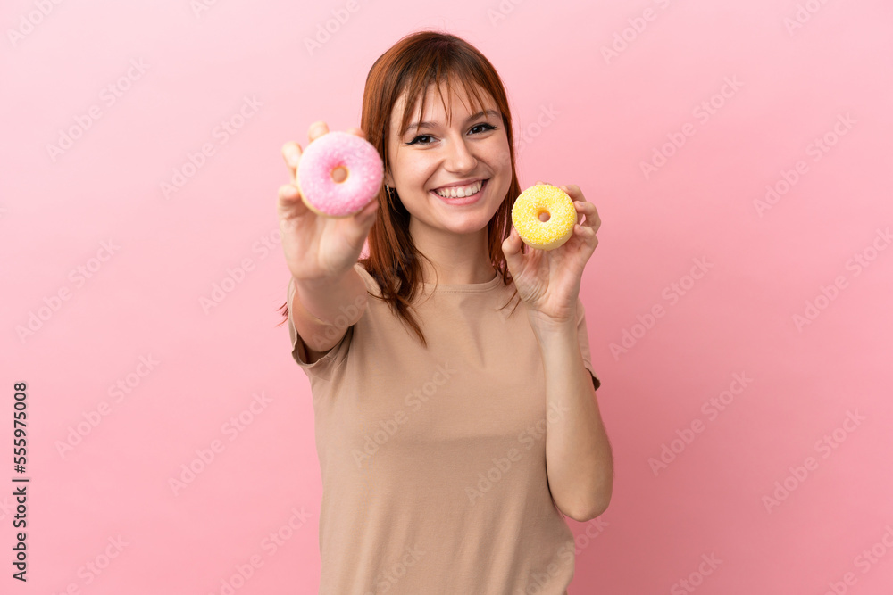 Redhead girl isolated on pink background holding donuts with happy expression Redhead girl isolated on pink background holding donuts with happy expression
