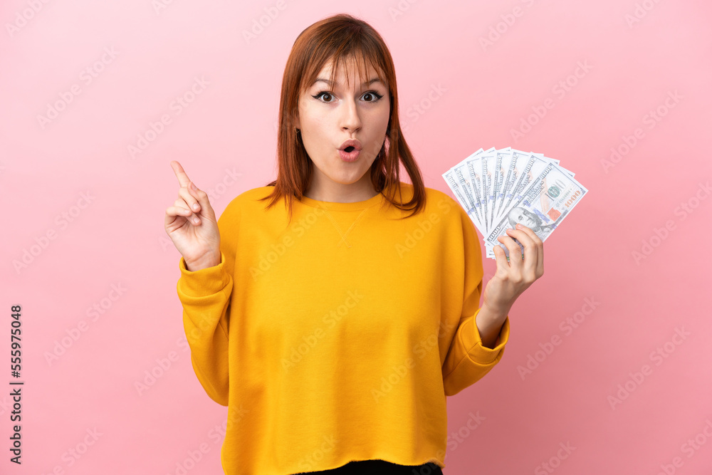 Redhead girl taking a lot of money isolated on pink background intending to realizes the solution while lifting a finger up Redhead girl taking a lot of money isolated on pink background intending to realizes the solution while lifting a finger up