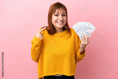 Redhead girl taking a lot of money isolated on pink background giving a thumbs up gesture