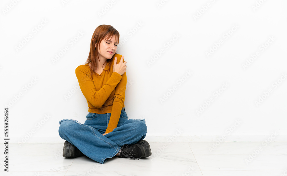 Redhead girl sitting on the floor isolated on white background suffering from pain in shoulder for having made an effort