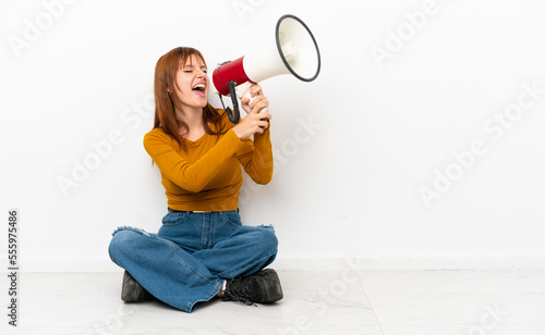 Redhead girl sitting on the floor isolated on white background shouting through a megaphone