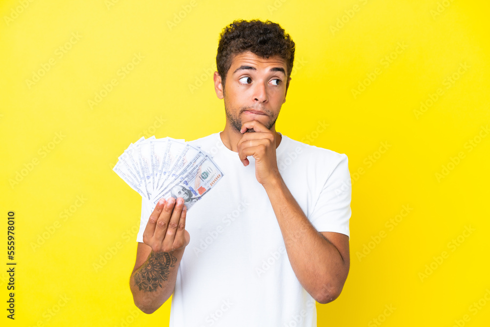 Young brazilian man taking a lot of money isolated on yellow background having doubts and thinking Young brazilian man taking a lot of money isolated on yellow background having doubts and thinking
