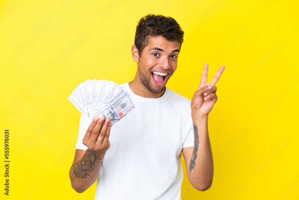 Young brazilian man taking a lot of money isolated on yellow background smiling and showing victory sign Young brazilian man taking a lot of money isolated on yellow background smiling and showing victory sign