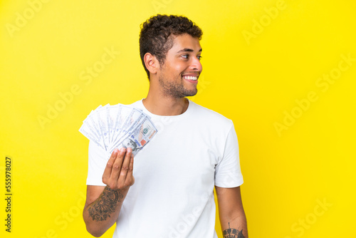 Young brazilian man taking a lot of money isolated on yellow background looking to the side and smiling